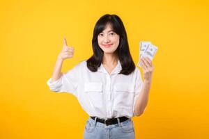 Young happy woman of Asian ethnicity wear white shirt and denim jean holding cash money in dollar and showing thumb up gesture against yellow background. Good investment and financial concept. photo