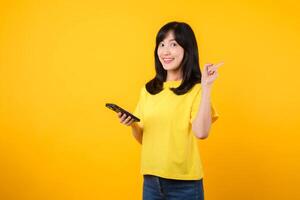 Capture young Asian woman wearing a yellow t-shirt and denim jeans points with finger hand gesture to free copy space while using smartphone. Perfect for illustrating the concept of app functionality. photo