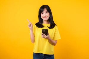 Capture young Asian woman wearing a yellow t-shirt and denim jeans points with finger hand gesture to free copy space while using smartphone. Perfect for illustrating the concept of app functionality. photo