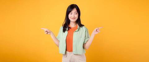 best deal promotion concept featuring young Asian woman 30s, wearing green shirt on orange shirt. With happy face, points her finger to free copy space, choose deals offers against yellow backdrop. photo