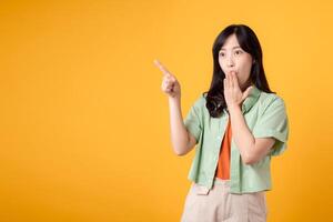 Capture engagement and excitement with Asian young woman wears pastel green shirt on orange shirt show happy smile while placing hand over mouth and pointing with a finger to free copy space. photo