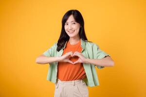 Experience the synergy of healthcare and wellness Asian young woman 30s, wearing a green shirt, showing heart hand gesture on her chest. Feel the essence of body wellness against yellow background. photo