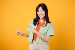 young Asian woman in her 30s wearing a green shirt on an orange shirt, pointing fingers to free copy space with enthusiasm. Explore the concept of discount shopping promotion with vibrant image. photo
