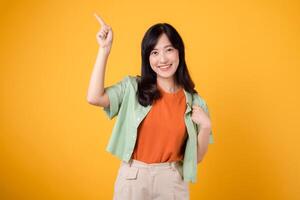 Unlock savings and shop now young Asian woman in her 30s, wearing a green shirt on an orange shirt. happy face and pointing finger to free copy space against vibrant yellow backdrop. photo