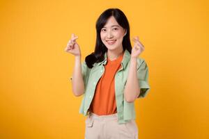 joven asiático mujer en su 30s vistiendo un naranja camisa y verde saltador graciosamente demostración mini corazón gesto y amable sonrisa, exudando afecto y sensibilidad. cuerpo idioma concepto. foto
