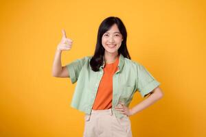 young Asian woman 30s, dressed in orange shirt and green jumper. Her thumbs up gesture, isolated on vibrant yellow background, embodies the concept of approval and positivity. photo