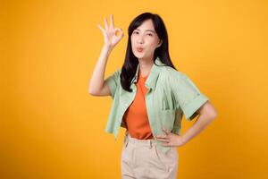 young Asian woman in her 30s, donning an orange shirt and green jumper. Her okay hand gesture and gentle smile, isolated on a vibrant yellow background, exude positivity through body language. photo