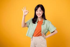 young Asian woman 30s, elegantly clad in orange shirt and green jumper. Her endearing okay hand gesture and gentle smile, set against a yellow background, reveal the beauty of body language. photo