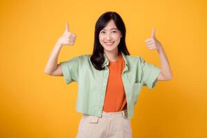 celebrar aprobación con joven asiático mujer 30, vistiendo en naranja camisa y verde puente. su pulgares arriba gesto, aislado en vibrante amarillo fondo, significa un concepto de positividad y acuerdo. foto