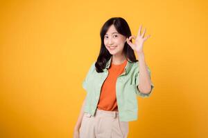 charming young Asian woman 30s, elegantly attired in orange shirt and green jumper. Her captivating okay hand gesture and gentle smile shine against a yellow background, body language concept. photo