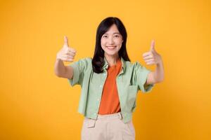 joven asiático mujer 30, esmeradamente ataviado en naranja camisa y verde puente. su pulgares arriba gesto, conjunto en contra un amarillo fondo, personifica el concepto de afirmación. foto