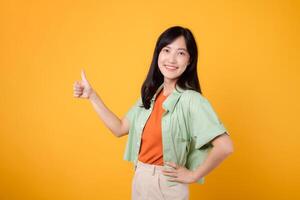 approval with cheerful young Asian woman 30s, elegantly dressed in orange shirt and green jumper. Her thumbs up gesture, set against vibrant yellow background, reflects the concept of encouragement. photo