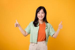 approval with cheerful young Asian woman 30s, elegantly dressed in orange shirt and green jumper. Her thumbs up gesture, set against vibrant yellow background, reflects the concept of encouragement. photo