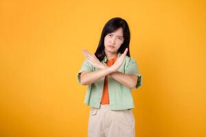 denial with a captivating young Asian woman 30s, donning an orange shirt and green jumper. Her cross hand gesture, isolated on a yellow background, represents the concept of refusal and rejection. photo