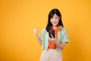 great news with a cheerful young Asian woman in her 30s, elegantly dressed in orange shirt and green jumper, raising a fist with smartphone on yellow studio background. Mobile concept photo