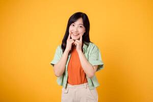 dental wellness with cheerful young Asian woman 30s, elegantly attired in orange shirt and green jumper points to her teeth on yellow background, emphasizing the significance of dental healthcare. photo