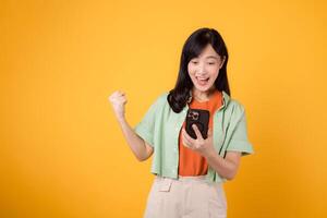el alegría de genial Noticias como un joven asiático mujer en su 30, poniéndose naranja camisa y verde saltador, celebra con un puño arriba gesto y teléfono inteligente en amarillo estudio antecedentes. móvil concepto. foto