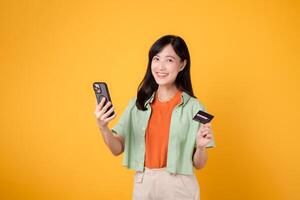 el alegría de en línea compras con un alegre joven asiático mujer en su 30, vistiendo naranja camisa y verde saltador, utilizando teléfono inteligente mientras participación crédito tarjeta en amarillo estudio antecedentes. móvil concepto. foto