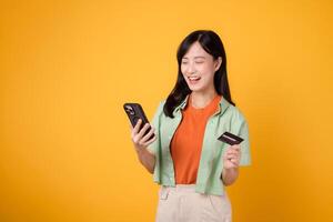 young 30s asian woman surprise face dressed in orange shirt and green jumper using smartphone while holding credit card isolated on yellow studio background. shopping online from mobile concept. photo
