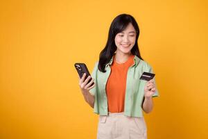 joy of online shopping with a cheerful young Asian woman in her 30s, wearing orange shirt and green jumper, using smartphone while holding credit card on yellow studio background. Mobile concept. photo