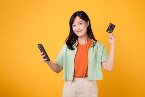 abrazo el facilitar de móvil compras con un encantador joven asiático mujer en su 30, vistiendo naranja camisa y verde saltador, utilizando teléfono inteligente mientras mostrando crédito tarjeta en amarillo estudio antecedentes foto
