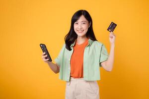 conveniencia de compras desde móvil con joven asiático mujer en su 30, esmeradamente vestido en naranja camisa y verde saltador, utilizando teléfono inteligente mientras exhibiendo crédito tarjeta en amarillo estudio antecedentes. foto