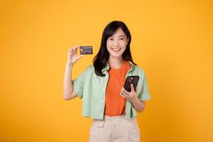 joven asiático mujer en su 30, vestido en naranja camisa y verde saltador, tiendas en línea utilizando teléfono inteligente con crédito tarjeta en amarillo estudio antecedentes. móvil concepto. foto