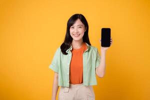 young Asian woman in her 30s, wearing orange shirt and green jumper, showcases smartphone screen display on yellow studio background. New mobile application concept. photo