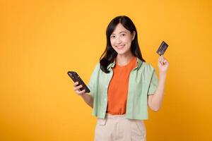 emoción de en línea compras con un alegre joven asiático mujer 30, vistiendo naranja camisa y verde saltador, utilizando teléfono inteligente mientras mostrando crédito tarjeta en amarillo estudio antecedentes. móvil concepto. foto