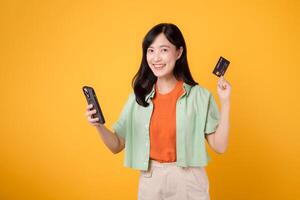 thrill of shopping from mobile with a captivating young Asian woman in her 30s, elegantly dressed in orange shirt and green jumper, using smartphone to show credit card on yellow studio background. photo