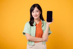 contento joven asiático mujer 30, vistiendo naranja camisa y verde saltador, muestra teléfono inteligente pantalla en amarillo estudio antecedentes. nuevo móvil solicitud, en línea compras, y nuevo móvil dispositivo concepto. foto