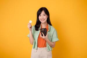 futuro de Finanzas con un encantador joven asiático mujer en su 30, vistiendo naranja camisa y verde saltador, utilizando teléfono inteligente y cripto moneda moneda en amarillo antecedentes. foto