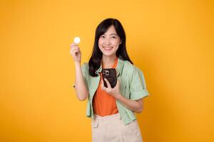 Happy young Asian woman in her 30s, wearing orange shirt and green jumper, showcases smartphone screen display and crypto currency coin on yellow background. Future finance concept. photo