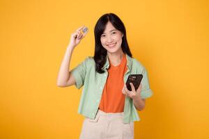 futuro Finanzas con joven asiático mujer en su 30, vestido en naranja camisa y verde saltador, exhibiendo cripto moneda moneda mientras participación teléfono inteligente en amarillo antecedentes. futuro Finanzas concepto. foto