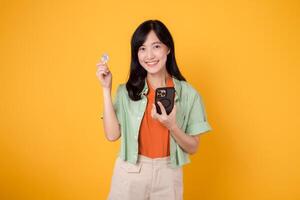 future of finance with cheerful young Asian woman in her 30s, donning orange shirt and green jumper, displaying smartphone screen and crypto currency coin on yellow background. Future finance concept. photo