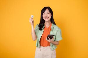 Happy young Asian woman in her 30s, wearing orange shirt and green jumper, showcases crypto currency coin while holding smartphone on yellow background. Future finance concept. photo