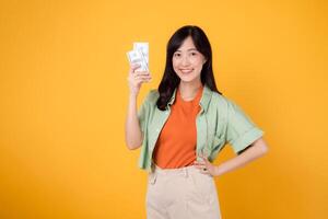 Happy young Asian woman in her 30s, wearing orange shirt and green jumper, showcases dollar currency while striking an akimbo gesture on yellow background. Financial money concept. photo