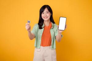 future finance with a vibrant young Asian woman in her 30s, dressed in orange shirt and green jumper, displaying smartphone screen display and crypto currency coin on yellow background. photo