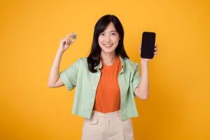 the future of finance with a captivating young Asian woman in her 30s, elegantly dressed in orange shirt and green jumper, using smartphone screen and crypto currency coin on yellow background. photo