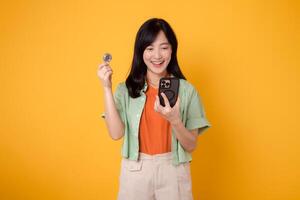 innovation of future finance with a vibrant young Asian woman in her 30s, dressed in orange shirt and green jumper, showcasing smartphone screen and crypto currency coin on yellow background photo