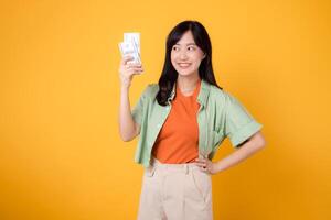 financial money with cheerful young Asian woman 30s, donning orange shirt and green jumper, displaying dollar currency while striking an akimbo gesture on yellow background. Financial money concept. photo