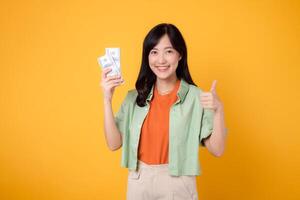 young 30s asian woman happy face dressed in orange shirt and green jumper showing dollar currency while showing thumb up hand sign gesture isolated on yellow background. finance business concept. photo