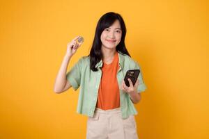 Embrace the future of finance with young Asian woman 30s, wearing orange shirt and green jumper, revealing crypto currency coin while holding smartphone on yellow background. Future finance concept. photo