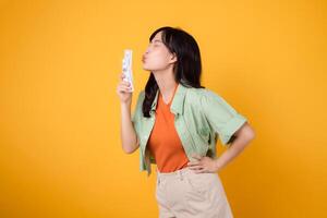 financial money with cheerful young Asian woman 30s, donning orange shirt and green jumper, kissing dollar currency while striking an akimbo gesture on yellow background. Financial money concept. photo