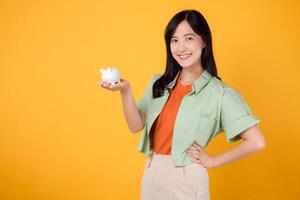 financial money with cheerful young Asian woman in her 30s, donning orange shirt and green jumper, displaying piggy bank while striking akimbo gesture on yellow background. Financial money concept. photo