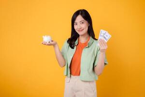 joven asiático mujer en su 30, vistiendo naranja camisa y verde saltador, demostración dólar moneda y cerdito banco en amarillo antecedentes. financiero dinero concepto. foto