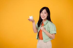 young Asian woman in her 30s, dressed in orange shirt and green jumper, showcasing piggy bank while pointing finger to free copy space on yellow background. Financial money concept. photo