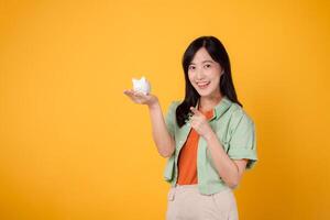 young Asian woman in her 30s, elegantly dressed in orange shirt and green jumper, revealing piggy bank while pointing finger to free copy space on yellow background. Financial money concept. photo