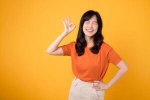 Expressing positivity, Asian woman 30s wears orange shirt, exhibits okay sign on vibrant yellow background. Hands gesture concept. photo