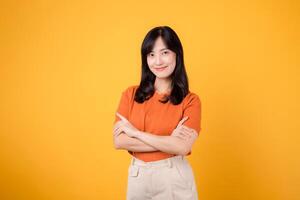 Embrace the confident vibes with a young Asian woman in her 30s, wearing an orange shirt, showcasing crossed arm sign gesture on yellow background. photo